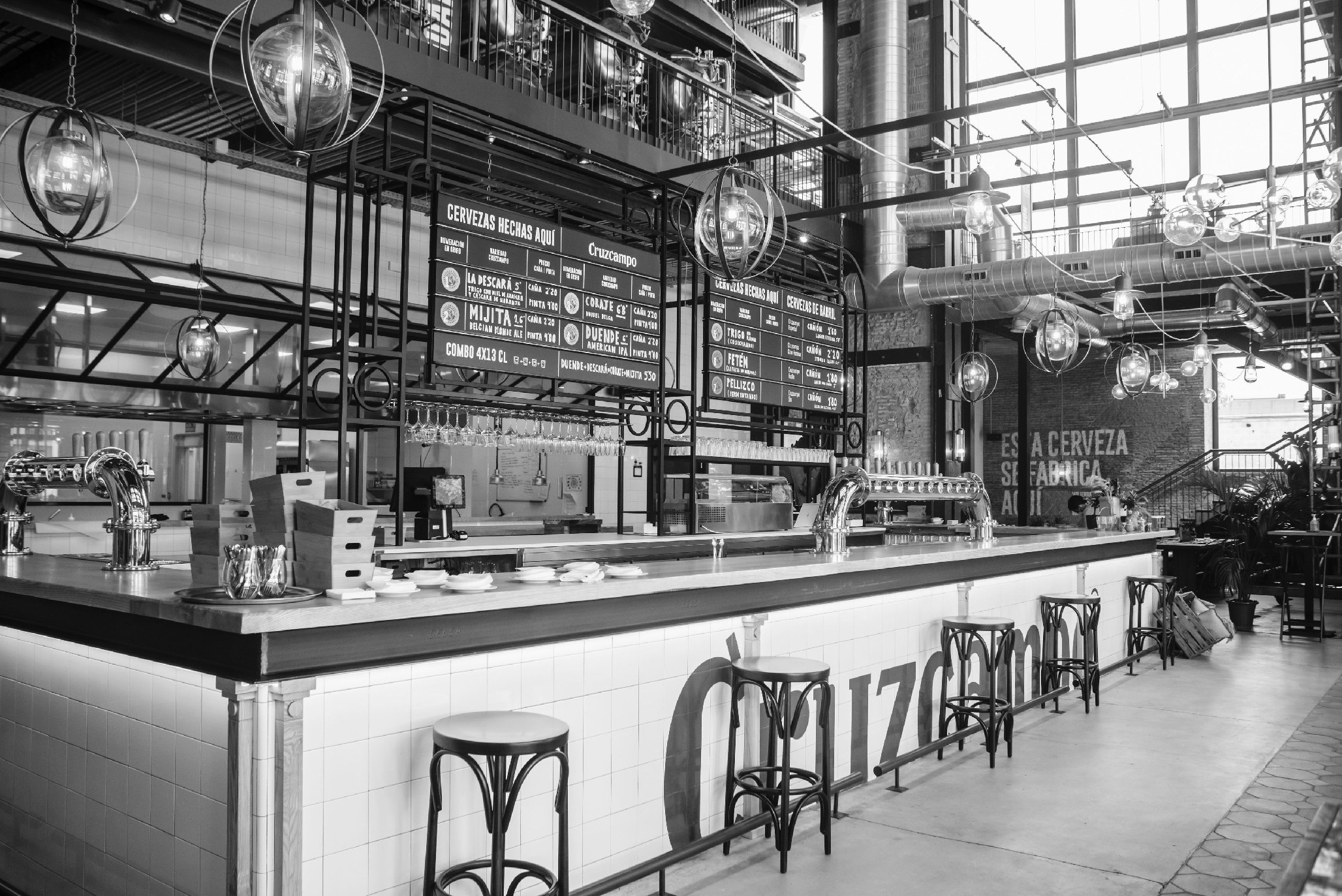 Fábrica de Cultura Cruzcampo en Sevilla: barra larga con el wordmark Cruzcampo, lámparas industriales suspendidas y pizarras de cervezas al fondo.